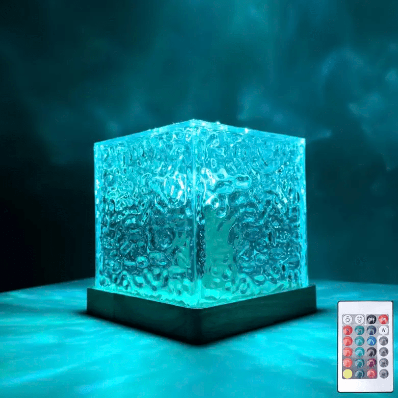 Textured clear cube lamp on a wooden base glowing teal-blue in a dark room, with a small remote control beside it. 1