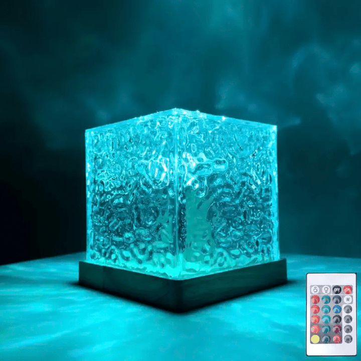 Textured clear cube lamp on a wooden base glowing teal-blue in a dark room, with a small remote control beside it. 1