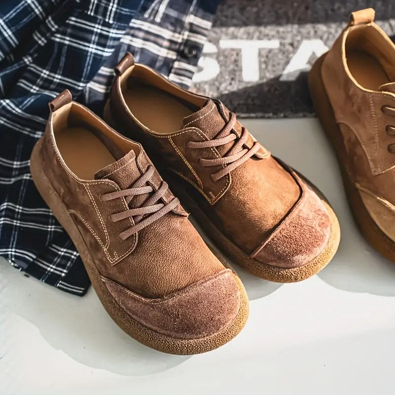 Brown and tan leather lace-up shoes with visible stitching arranged on a white surface beside a navy plaid shirt.