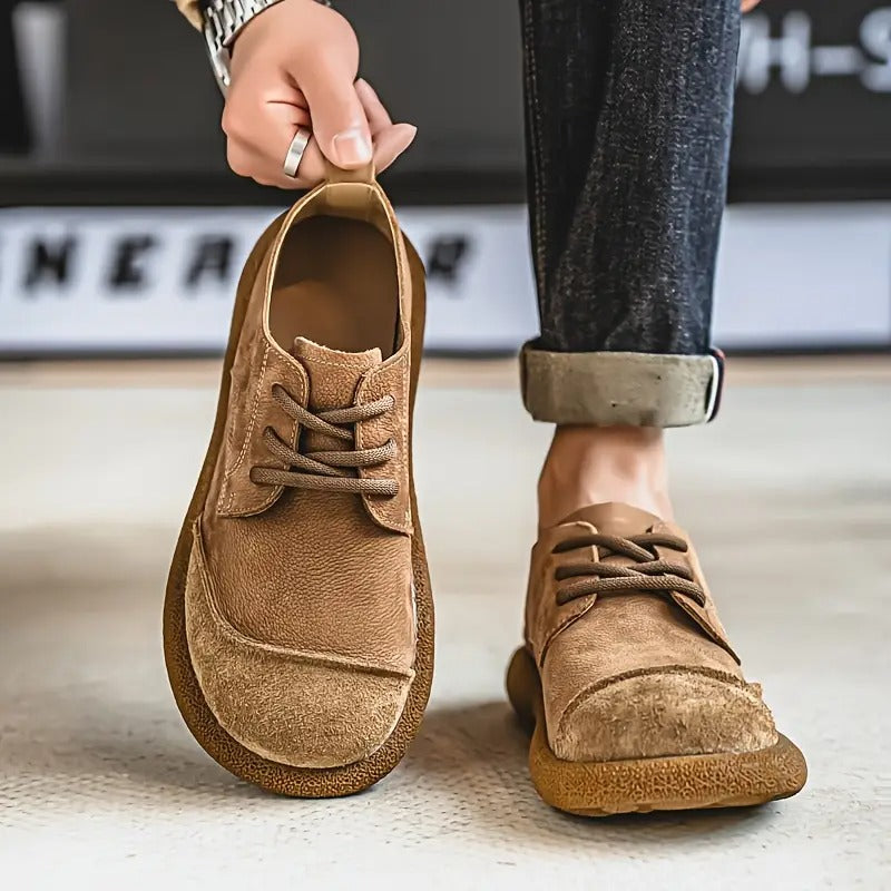 Brown leather lace-up shoes detail with one shoe held by the heel tab and one worn with rolled dark jeans.