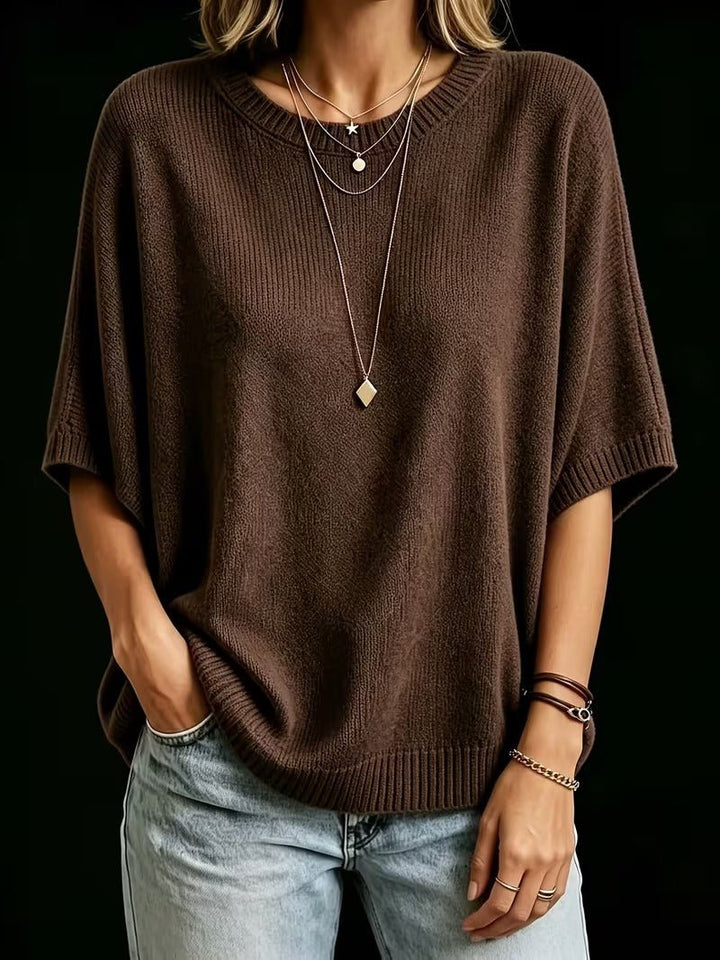 Dark brown knit batwing top on a model with one hand in a pocket, layered necklaces and bracelets.