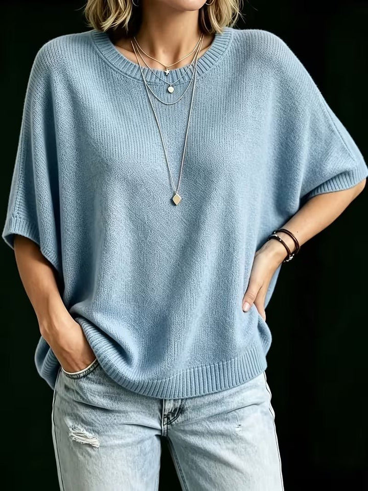 Light blue knit batwing top worn with one hand in a jeans pocket and the other on the hip.