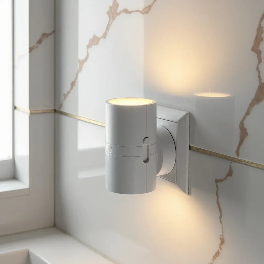 White cylindrical plug-in lamp lit in an outlet beside a white marble wall with gold trim.
