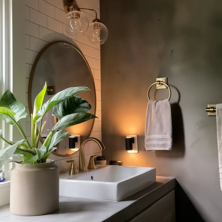 Another view of two black plug-in lamps glowing beside a bathroom mirror with a brass faucet and towel ring.