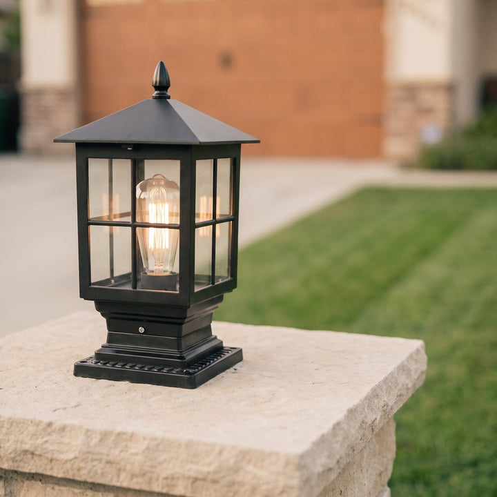 Glowing black post lamp on a light stone pillar in daylight, with a green lawn and a driveway blurred behind. 3