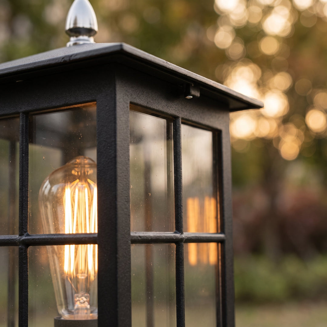 Close-up of the lamp's textured black frame, glass panes, silver-toned finial and glowing filament bulb, with soft golden light blurred behind. 5