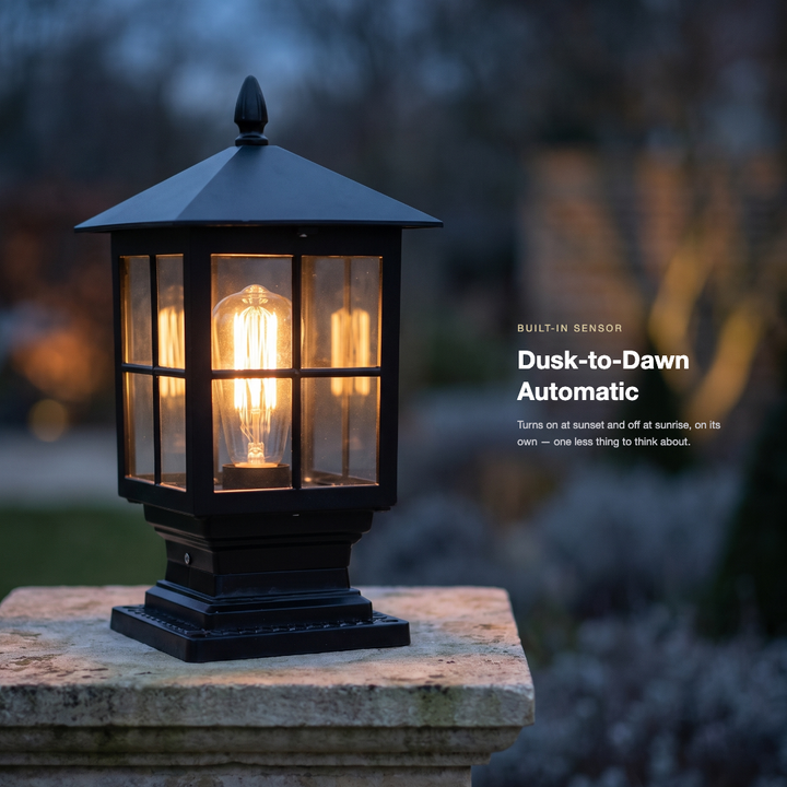 Lit black pillar lamp on a weathered stone post at dusk, with a blurred garden behind and a caption about automatic dusk-to-dawn operation. 2