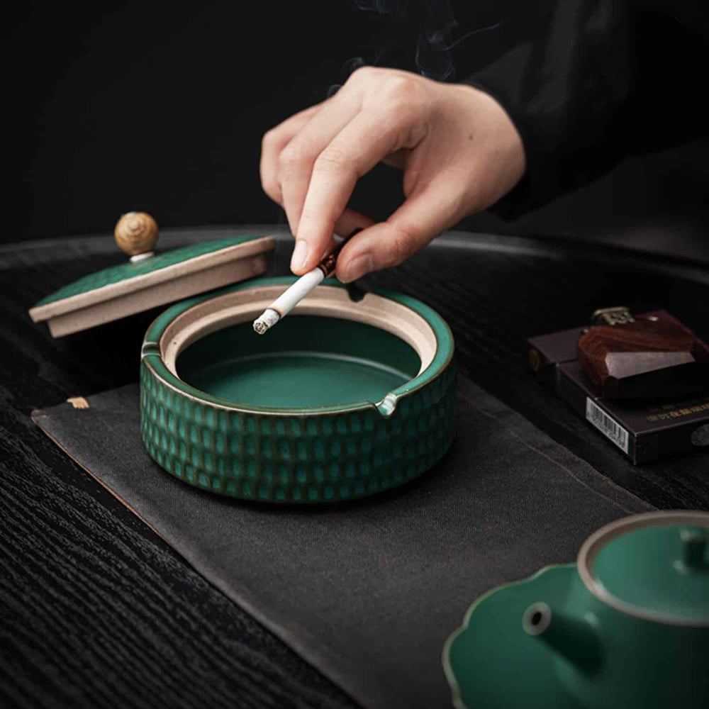 Green ceramic ashtray detail with the lid beside it and a lit cigarette held above an open bowl.