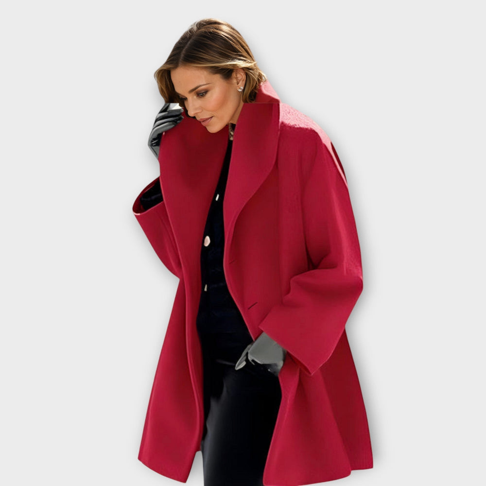 Model in the same wide-lapel coat in a deep raspberry red, worn open over black with gray gloves, looking down. 2