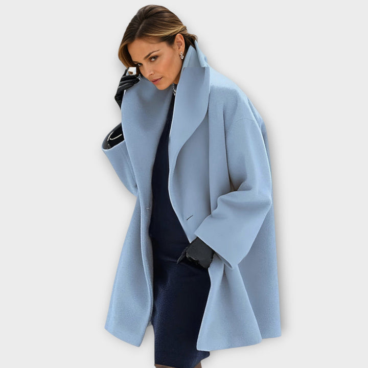 Woman wearing the oversized wide-lapel coat in light powder blue over a navy dress, with black leather gloves and a hand raised. 1