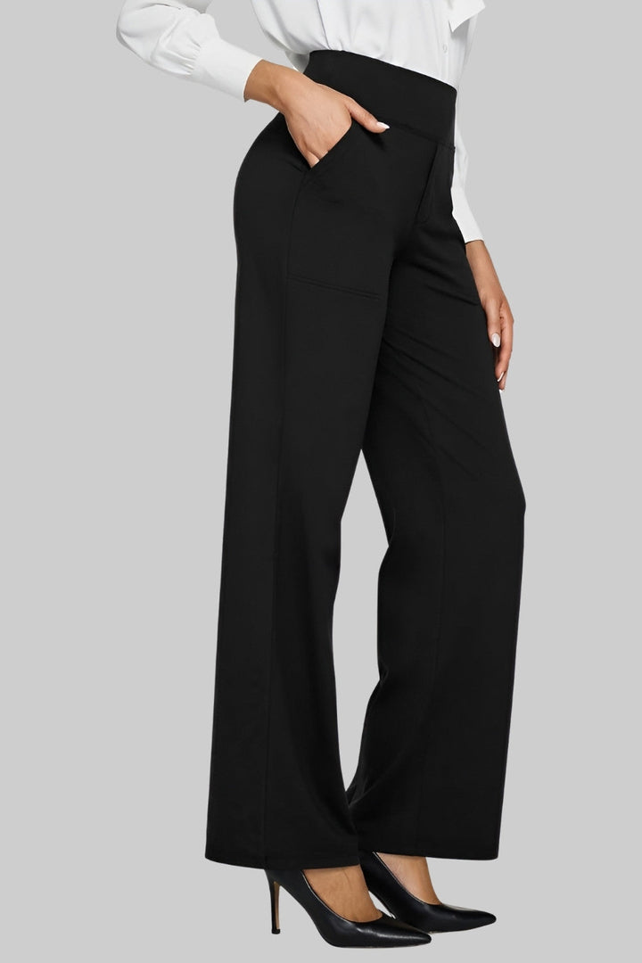 Women's black high-waist pants styled for the office with a white button-up shirt and black pointed high heels, side view with hand in pocket.