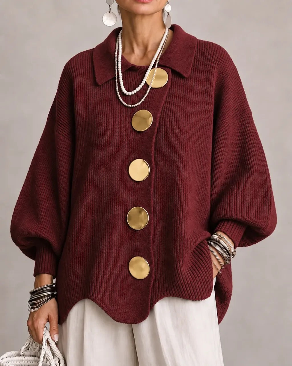 Burgundy ribbed cardigan on a model with large gold buttons, pointed collar and wavy hem over white pants. 19