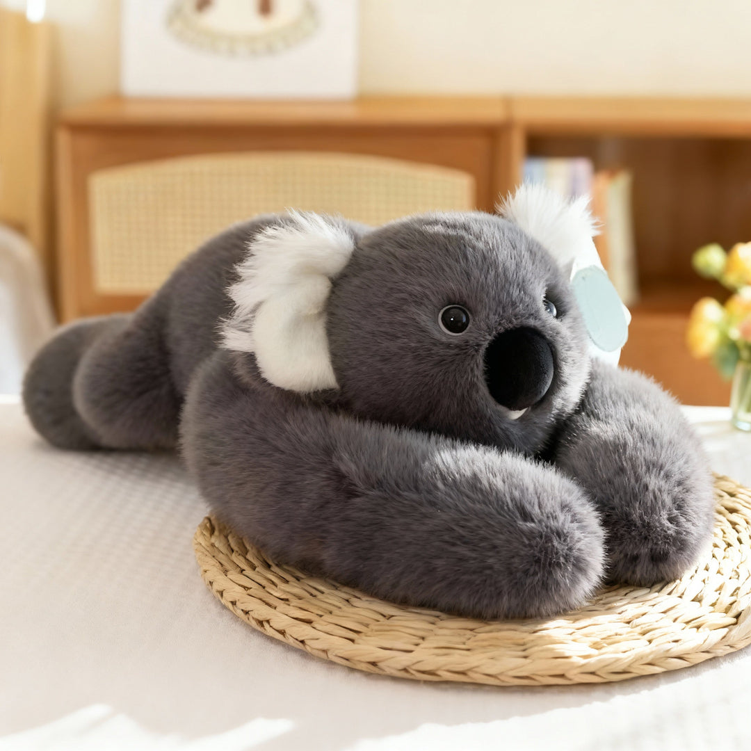 Gray koala plush with fluffy white ears and a large black nose lying on a woven straw mat with long arms curled around. 16