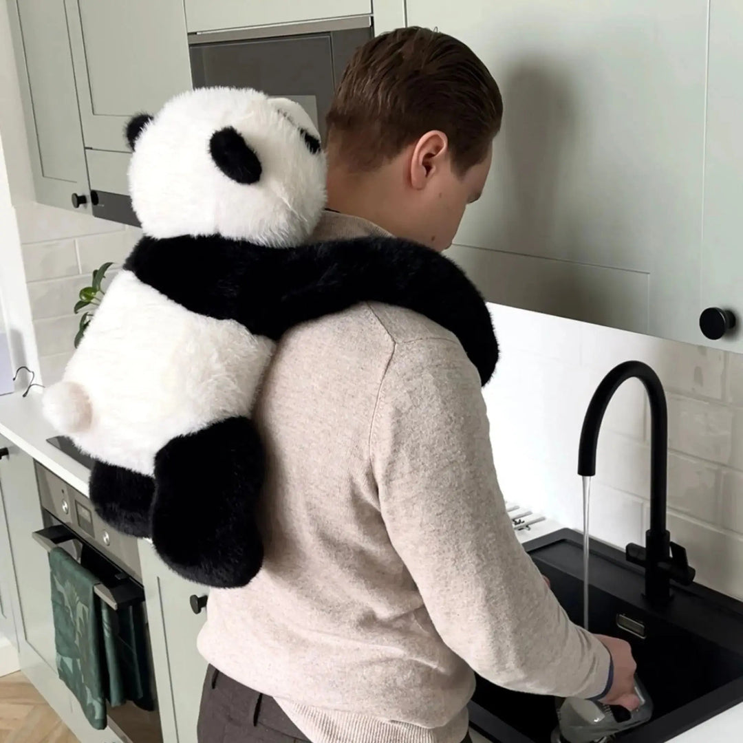 Man washing dishes at a kitchen sink with the panda plush wrapped around his shoulders and hanging down his back. 10