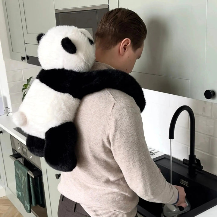 Man washing dishes at a kitchen sink with the panda plush wrapped around his shoulders and hanging down his back. 10