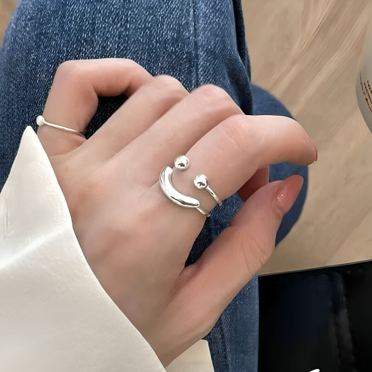 Silver open-band smiley ring with two ball ends worn on a model's index finger, shown with a white sleeve and denim. 1