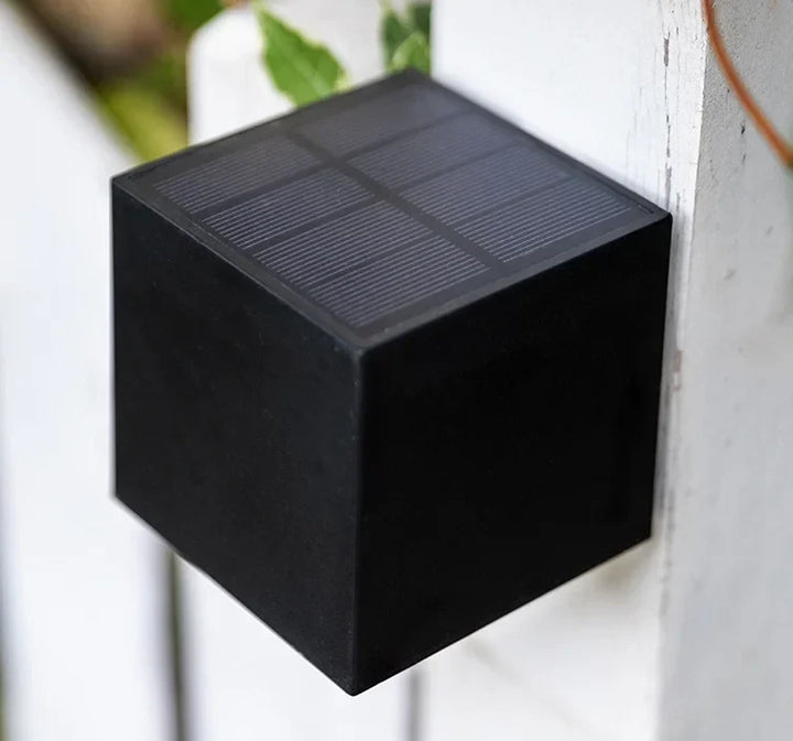Black solar wall light detail with a solar panel on top, attached to a white painted wooden post outdoors.