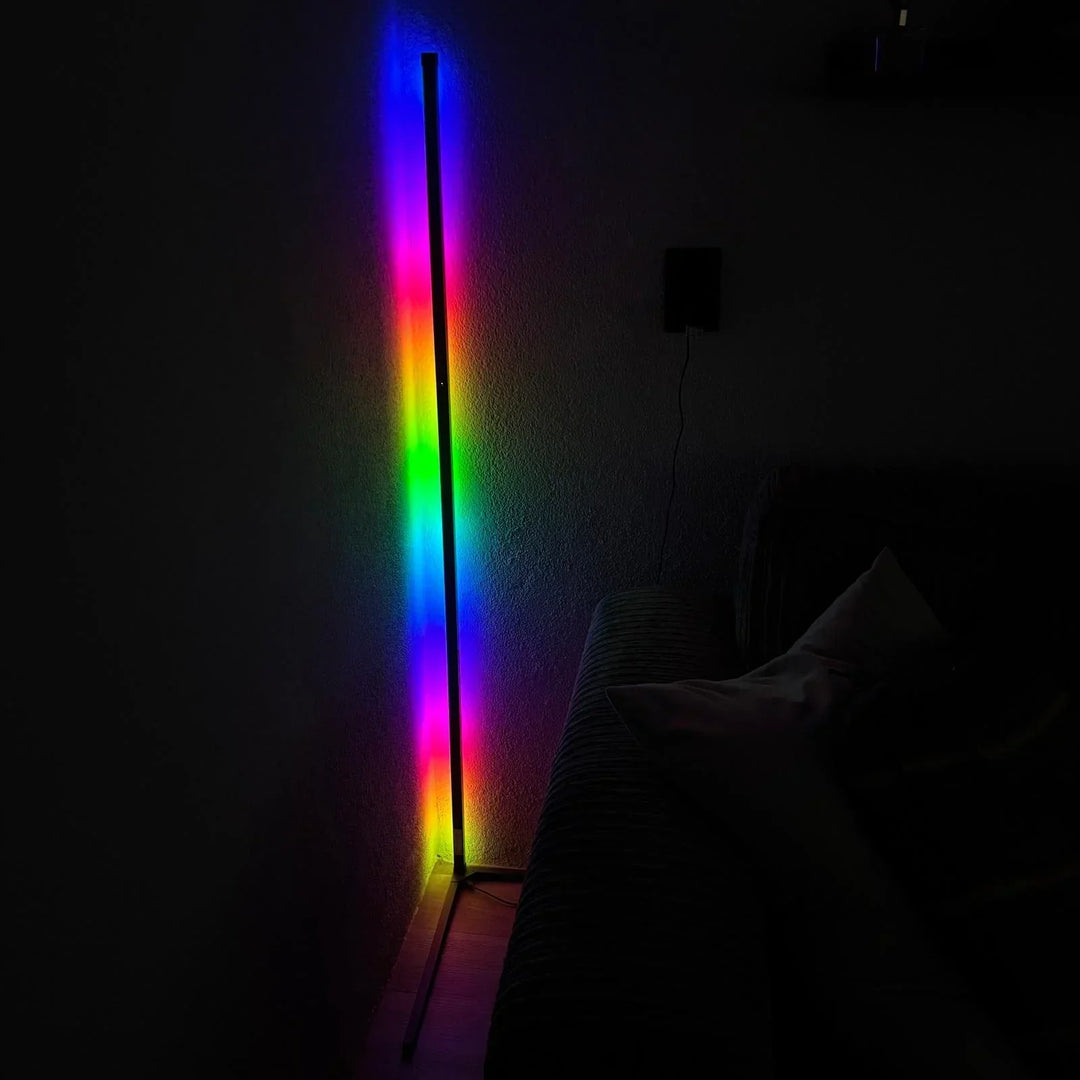 Corner lamp glowing in a rainbow of colors on a dark wall with a sofa nearby.