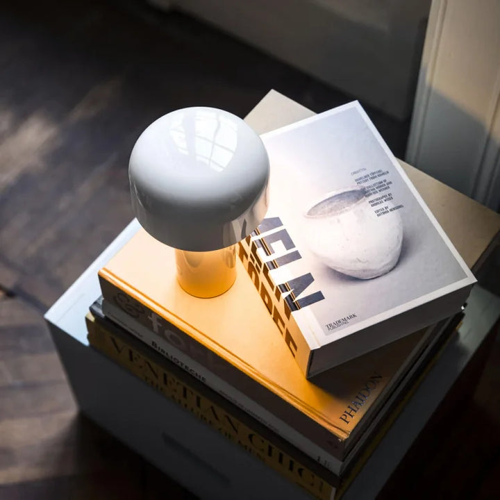White glossy lamp seen from above, glowing warmly on a stack of books on a small cabinet 20