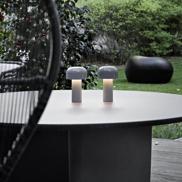 Two small gray lamps lit on a round outdoor table with a garden behind 23