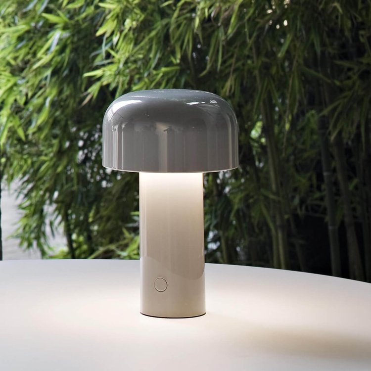 Gray mushroom lamp lit on a white table with green bamboo plants in the background 9
