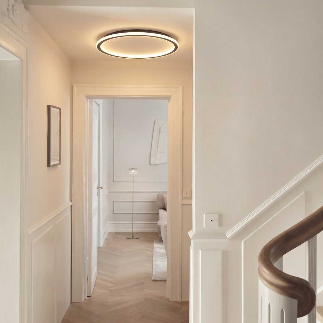 Glowing ring ceiling light in a bright hallway with white paneled walls, herringbone floor and a curved wooden stair railing. 5