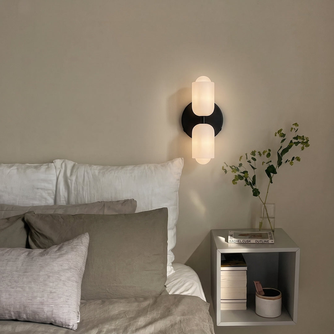 Black wall light glowing above a bedside shelf with books and a vase of green stems, next to a bed with gray and white linens. 5