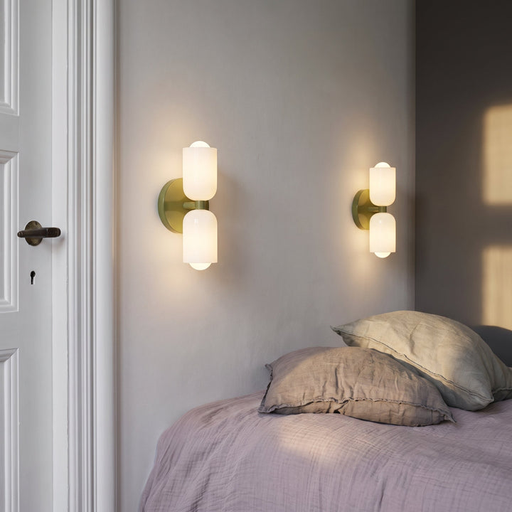 Two lit wall lights with olive green backplates and white glass shades on a gray bedroom wall above a bed with linen pillows. 4