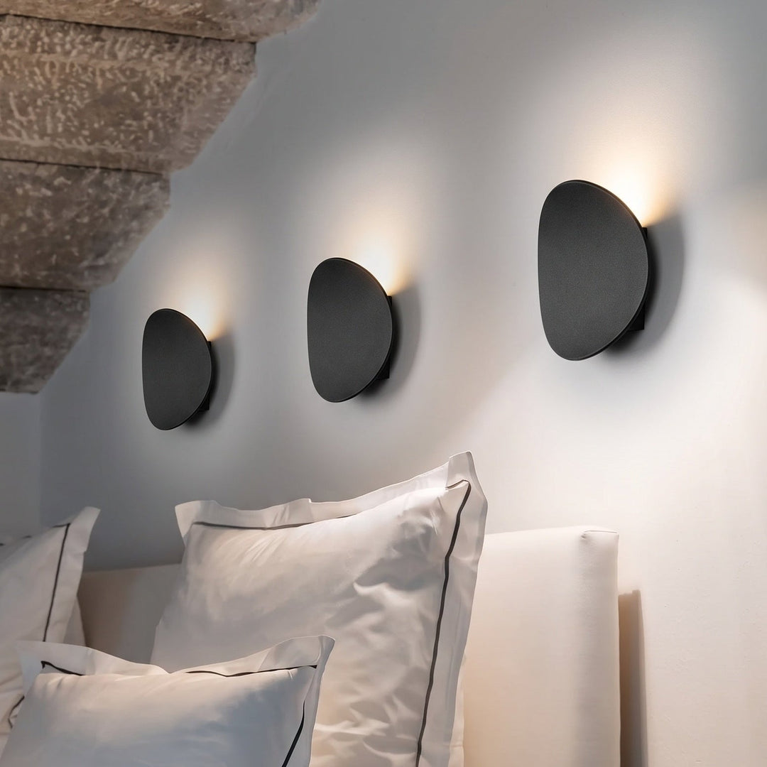 Three black pebble-shaped wall lamps in a row above a bed with white pillows, casting warm light upward onto the wall. 1