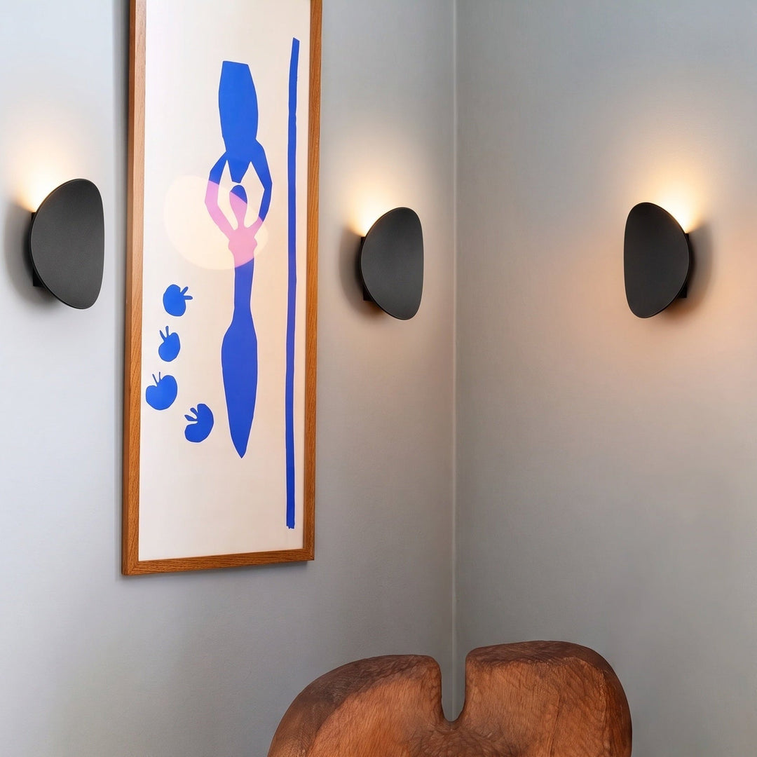Three black oval wall lamps glowing upward around a framed blue abstract print and a carved wooden stool in a corner. 2