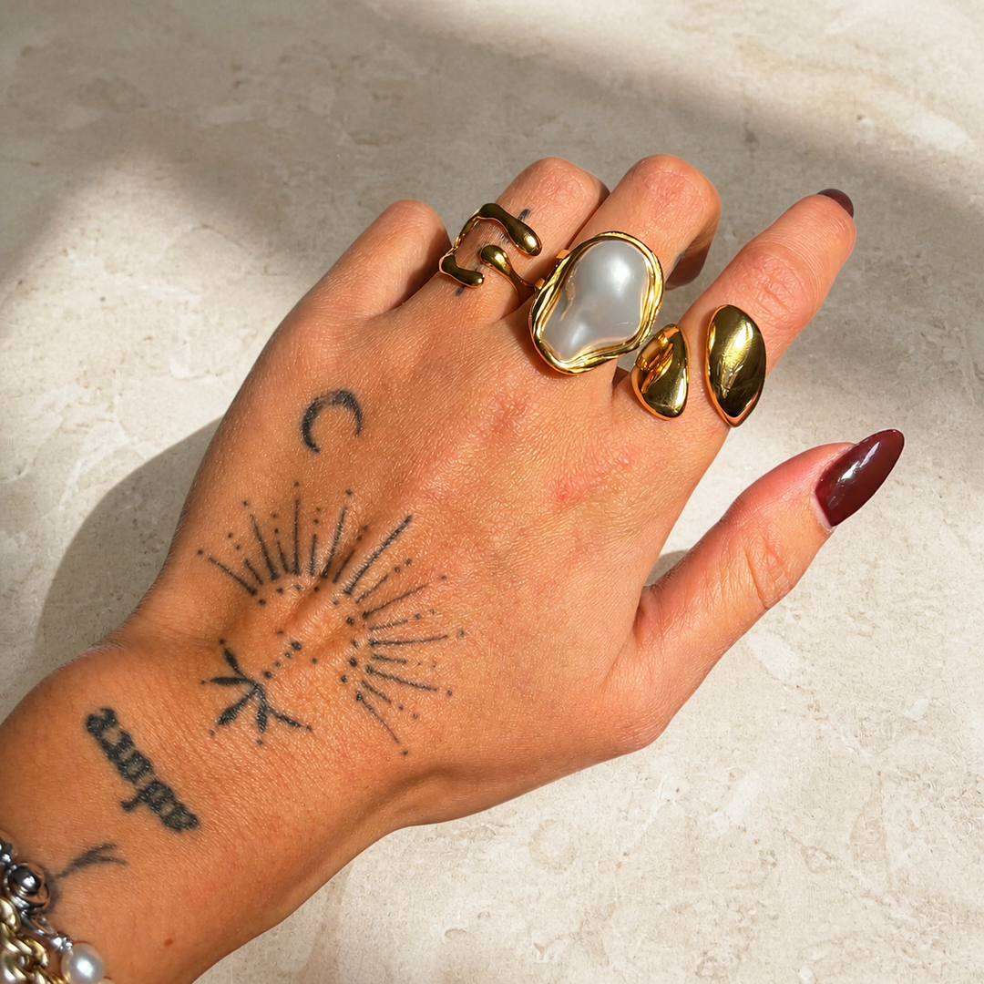 Tattooed hand resting on pale stone, wearing the gold open ring beside a large pearl-style ring and another wavy gold ring. 4