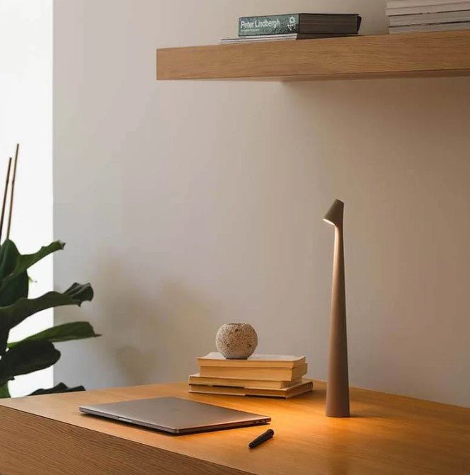 Slim tapered beige table lamp glowing warmly on a wooden desk beside a closed laptop, stacked books and a stone ornament. 21