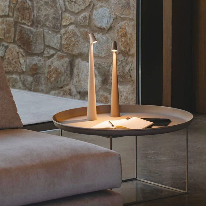 Another view of two beige rechargeable LED table lamps glowing on a tray table beside an armchair.