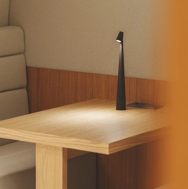 Another view of a black rechargeable LED table lamp lit on a light wooden table in a booth.