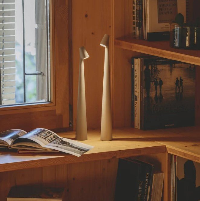 Another view of two taupe rechargeable LED table lamps lit on a pine wood shelf by a window.