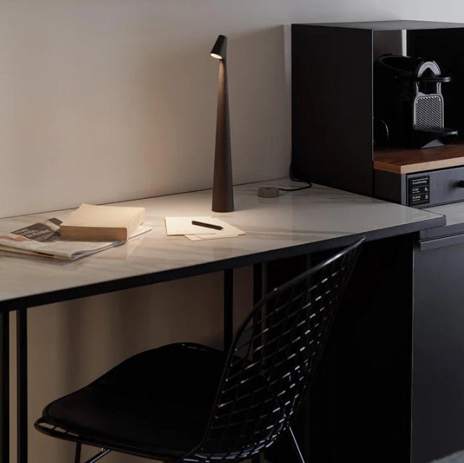 Another view of a dark brown rechargeable LED table lamp on a marble-top desk with a charging base.