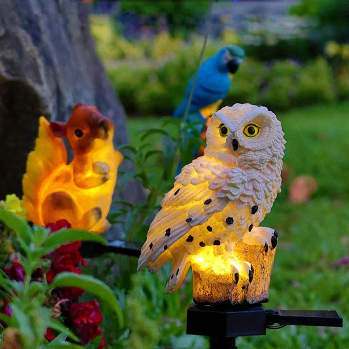 Glowing white owl light on a garden stake beside lit squirrel and blue parrot lights among red flowers.
