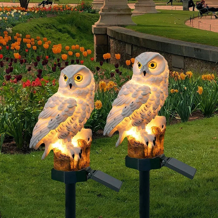 White owl solar lights glowing on stakes in a lawn with tulips and daffodils.