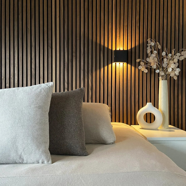 Black cube wall lamp lighting a walnut slat wall above a bed with cushions, a ring-shaped vase and dried branches. 5
