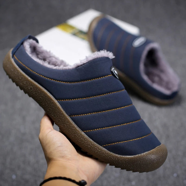 Hand holding a navy slipper up to show orange stitching, fleece interior and textured brown sole 6