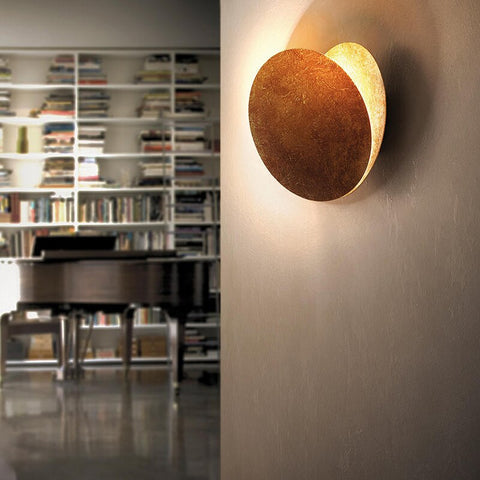 Side view of a tilted copper-toned disc lamp on a wall beside a bookshelf and piano 3