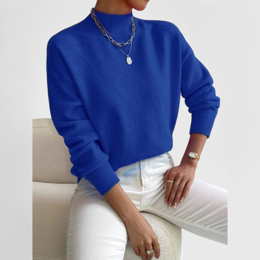 Women's sweater in royal blue with ribbed mock collar and cuffs, silver necklaces and white jeans.