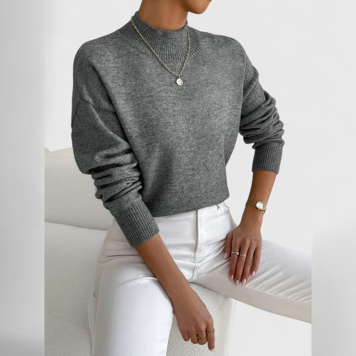 Women's sweater in heathered gray with a ribbed mock neck and cuffs, plus a pendant necklace and white jeans.