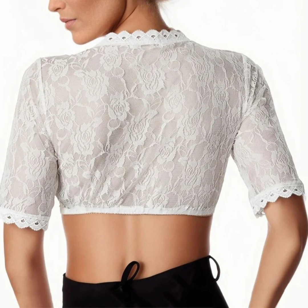 Back view of a white lace dirndl blouse with sheer rose-pattern fabric, a scalloped neckline and a cropped hem over black.