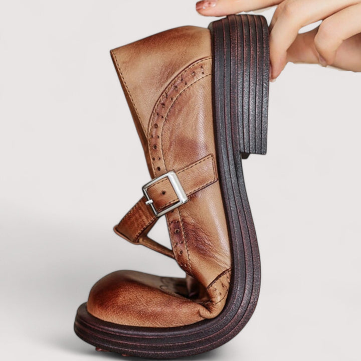 Hand holding a brown leather Mary Jane upright and flexed, showing the bendable sole, buckle strap and brogue perforations. 3