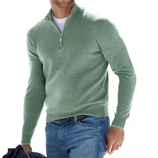 Man in a sage green quarter-zip knit sweater, worn over a white tee with blue jeans, holding a jacket.