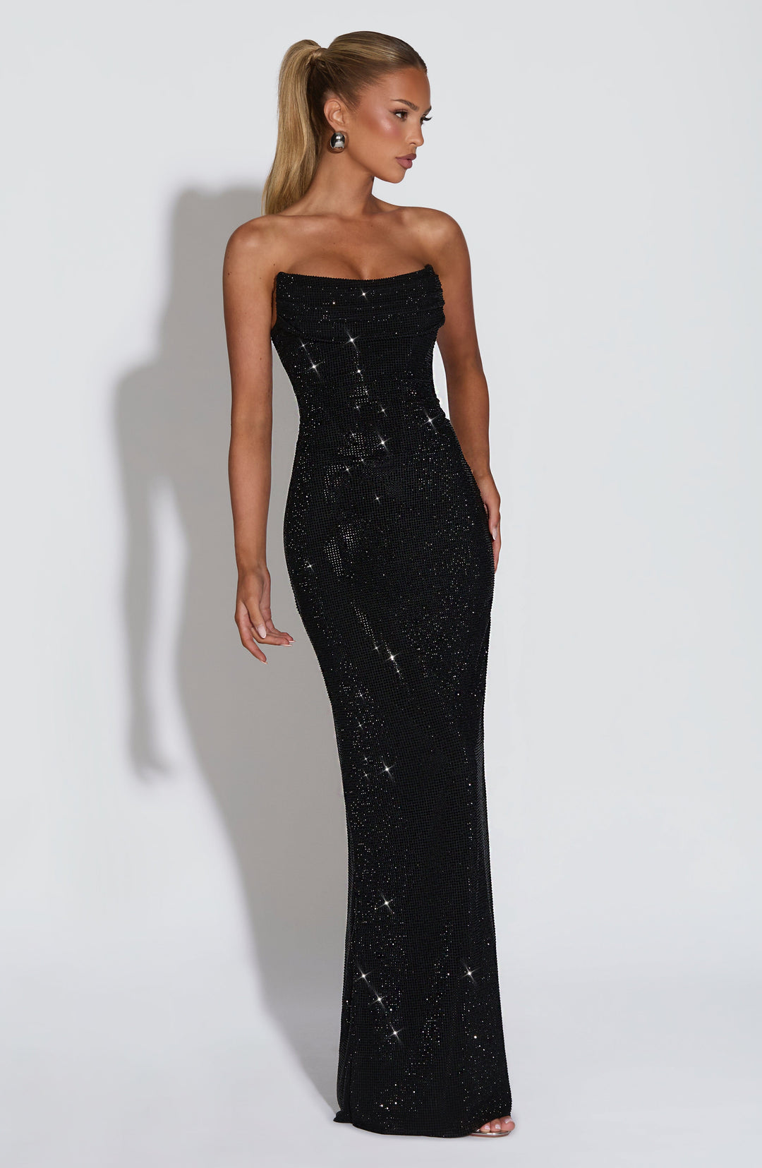 Three-quarter side view of the black rhinestone maxi dress on a model, showing its fitted full-length silhouette 25