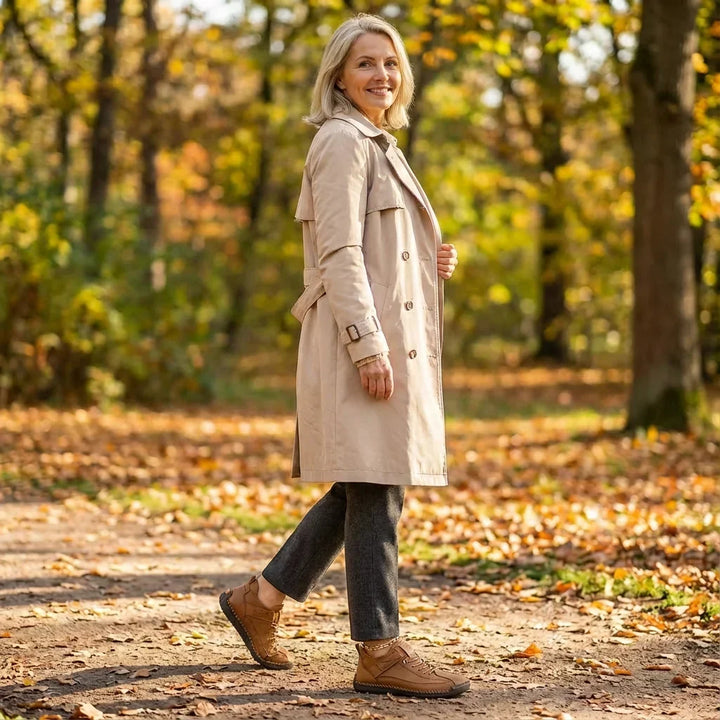 Smiling woman in a beige trench coat and gray trousers walks through an autumn park wearing brown sneakers. 11