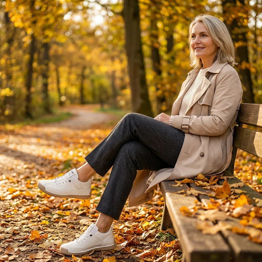 Woman in a trench coat sits on a park bench among fallen leaves, legs crossed, wearing white sneakers. 12