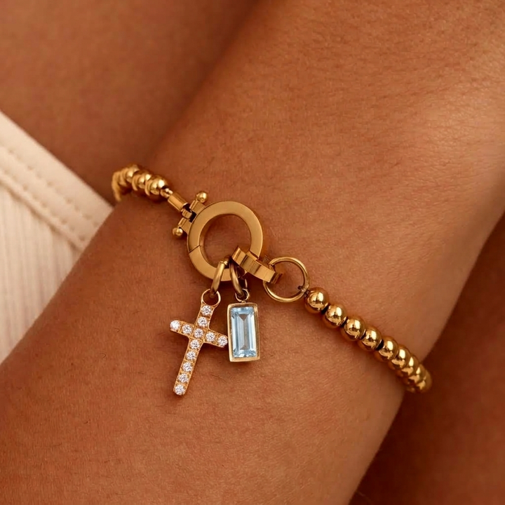 Gold-tone bead bracelet worn on a wrist with a crystal cross charm and light blue baguette stone charm.